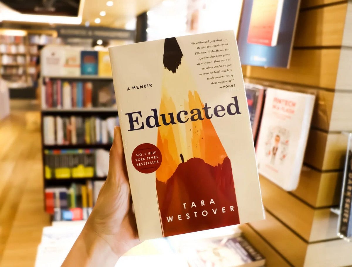 A person holding the book Educated by Tara Westover in a bookstore with blurred bookshelves in the background.